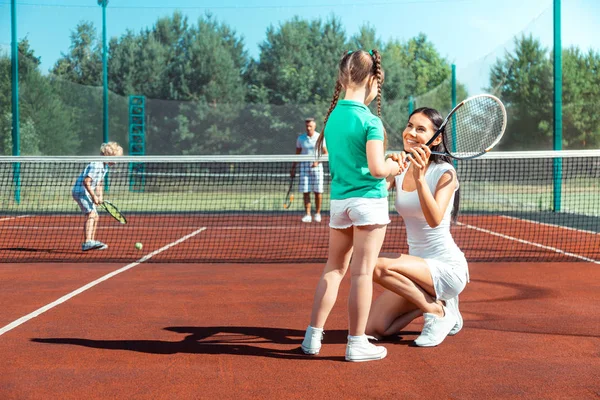 Mamãe de cabelos escuros sorrindo enquanto ensinava garota jogando tênis — Fotografia de Stock