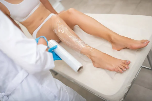 Cosmetologist covering leg of a client with plastic wrap. — Stock Photo, Image