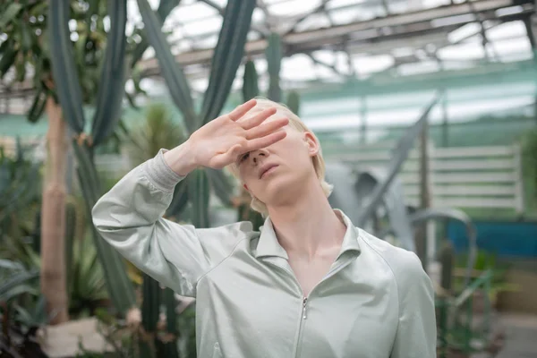 Blonde man hiding face while posing in greenhouse