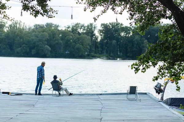 Hombre de pie cerca de padre sentado y pescando — Foto de Stock