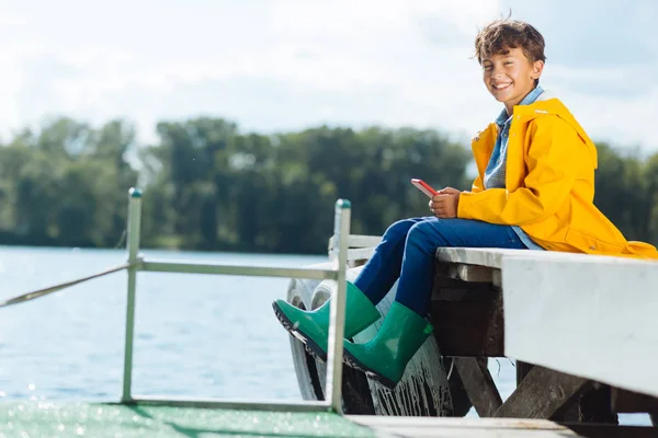 Pojke bär gröna regn stövlar leende när du sitter nära floden — Stockfoto