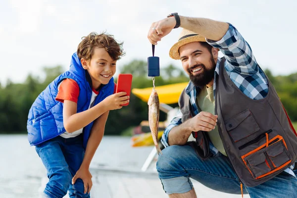 Cheerful boy taking picture of fish while fishing with dad