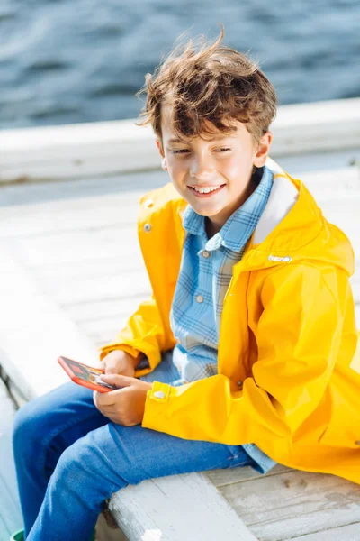 Aranyos mosolygó fiú viselt sárga esőkabát ülés közelében folyó — Stock Fotó