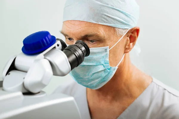 Poważny dojrzały lekarz siedzi przed mikroskopem — Zdjęcie stockowe