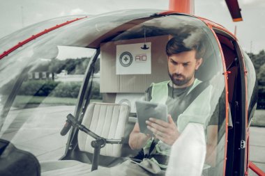 Küçük helikopterde otururken tabletini kullanan sakallı pilot