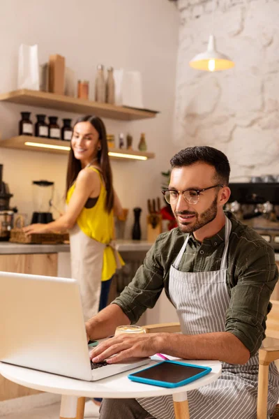 Marido trabalhando no laptop enquanto esposa fazendo café em sua cafetaria — Fotografia de Stock