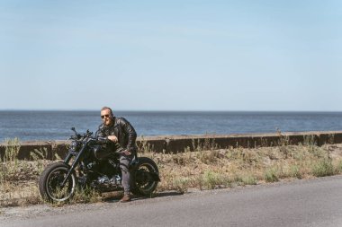 Deniz kenarında motosikletinin üstünde oturan yakışıklı, vahşi adam.