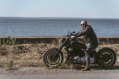Yakışıklı sakallı motosikletli deniz kenarında oturuyor.