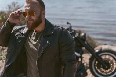 Bisikletinin yanında duran ve güneş gözlüğünü takan havalı bir motorcunun portresi.