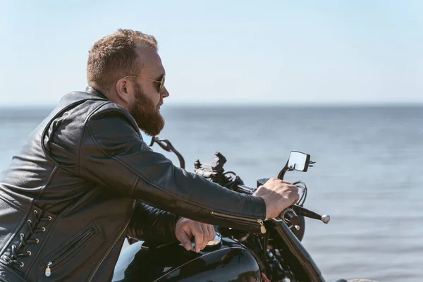 Вид збоку гарний бородатий байкер позує, сидячи на своєму мотоциклі — стокове фото