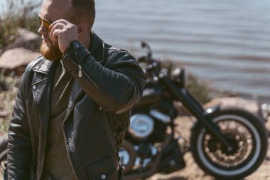 Motosikletinin yanında poz veren yakışıklı bir motorcunun portresi güneş gözlüğünü tamir ediyor.