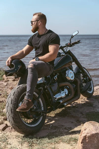 Bonito homem musculoso sentado em sua moto olhando para o mar — Fotografia de Stock
