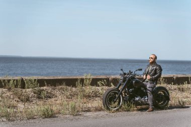 Deri ceketli seksi adam okyanusun kenarında bisiklette oturuyor.