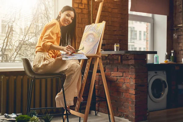 Radosna młoda kobieta maluje obraz na sztalugach w domu — Zdjęcie stockowe