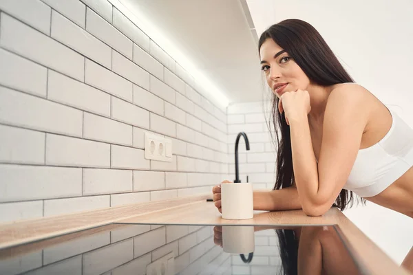 Улыбающаяся молодая женщина наклоняется над кухонной скамейкой, держа чашку напитка — стоковое фото