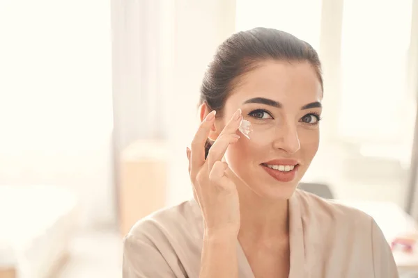 Nice-looking young woman moisturizing skin under her eyes — Stock Photo, Image