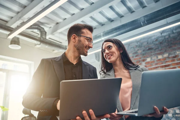 Compañeros sonrientes sosteniendo computadoras portátiles mientras discuten su proyecto —  Fotos de Stock