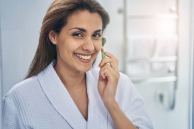 Güzel genç bayan banyoda yüz masajı aleti kullanıyor.