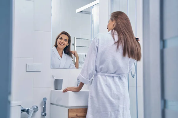Señora atractiva en albornoz mirando en el espejo en el baño —  Fotos de Stock