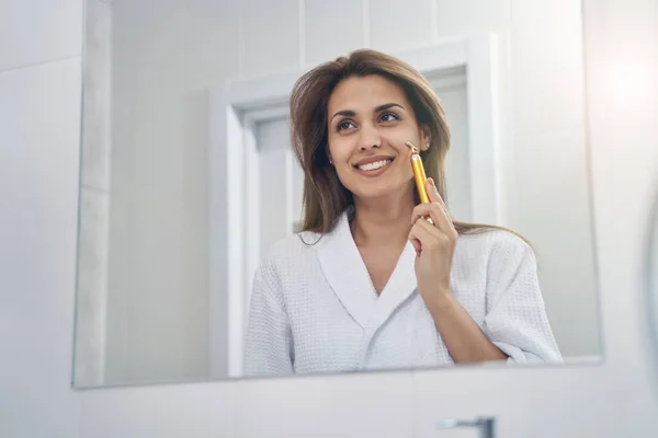 Vidám fiatal nő használ arany arc masszírozó a fürdőszobában — Stock Fotó