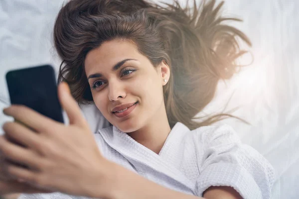 Encantadora joven acostada en la cama y haciendo selfie —  Fotos de Stock