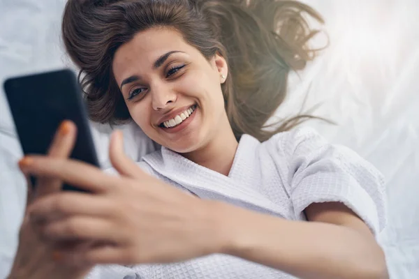 Весела молода жінка лежить в ліжку і використовує мобільний телефон — стокове фото