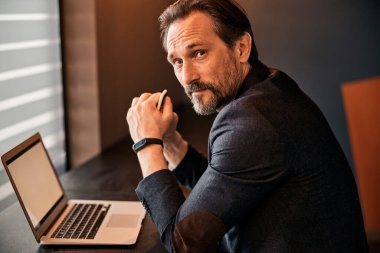 Siyah ceketli, sakallı, olgun iş adamı ofis masasında oturuyor.