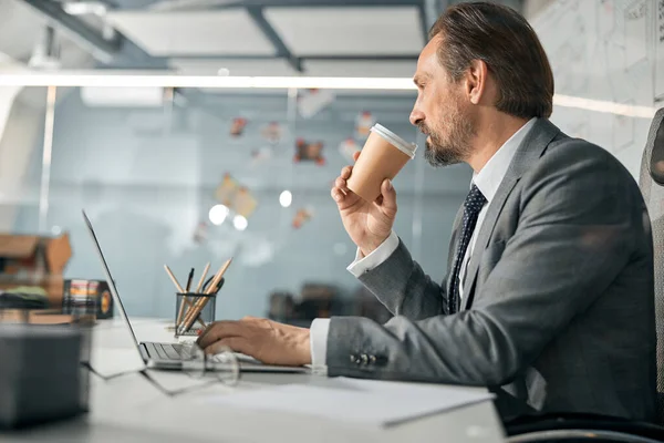 매력적 인 사업가가 컴퓨터를 사용하고 손에 커피를 들고 있는 모습 — 스톡 사진