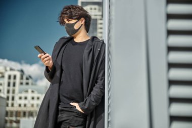 Açık havada akıllı telefon kullanan, tıbbi maske takan yakışıklı bir adam.