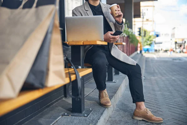 Стильный мужчина пьет кофе и пользуется ноутбуком в кафе на открытом воздухе — стоковое фото