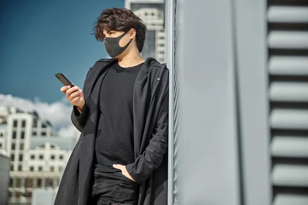 Chico guapo en mascarilla médica usando teléfono inteligente al aire libre —  Fotos de Stock