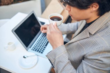 Genç adam kahve içiyor ve açık kafede dizüstü bilgisayar kullanıyor.