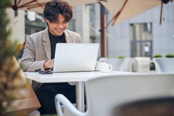 Χαρούμενος νεαρός που χρησιμοποιεί φορητό υπολογιστή σε υπαίθριο καφέ — Φωτογραφία Αρχείου