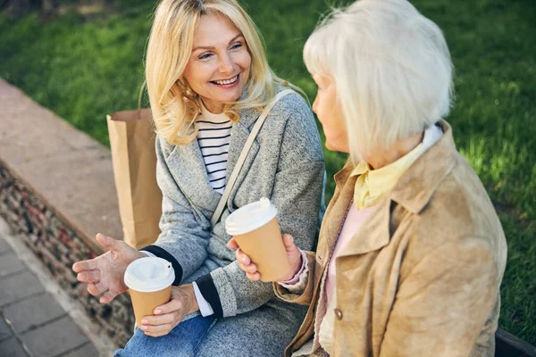 Duas mulheres bonitas conversando e bebendo café saboroso enquanto sentado no banco — Fotografia de Stock