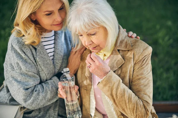 Пожилая женщина пьет воду, в то время как другая женщина сидит рядом с ней — стоковое фото