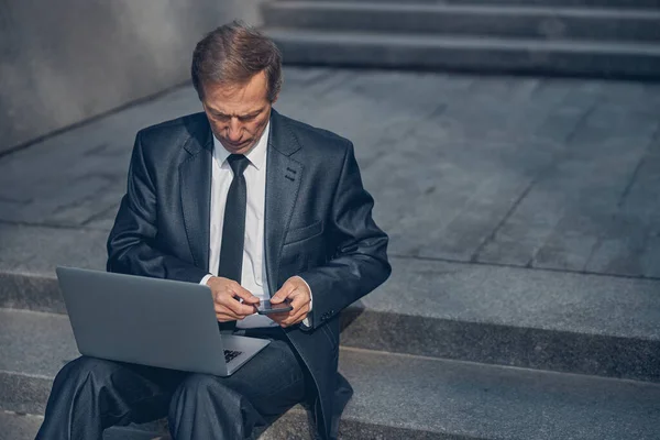 Allvarlig affärsman som använder bärbar dator och mobiltelefon på gatan — Stockfoto