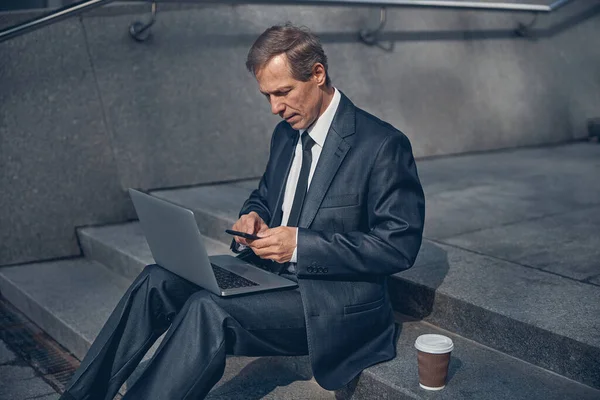 Красивый бизнесмен, использующий ноутбук и смартфон на улице — стоковое фото