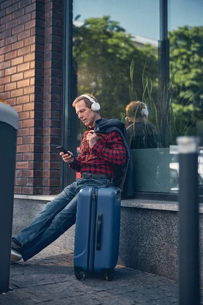 Knappe man met reiskoffer die buiten naar muziek luistert — Stockfoto