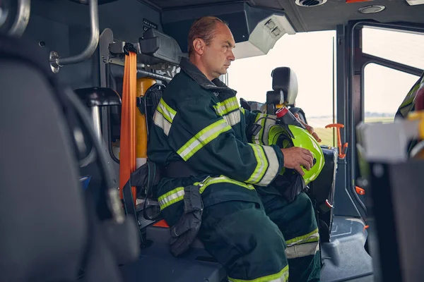 Bild på vuxen brandman med hjälm i händerna sittande i brandbil — Stockfoto