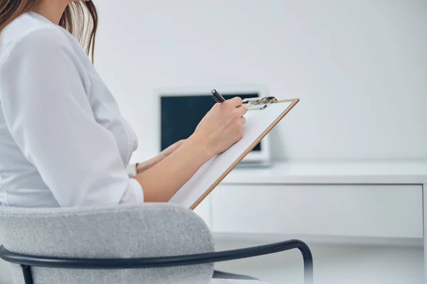 Woman in white medicine uniform sitting on the chair while writing notes — Stock Photo, Image