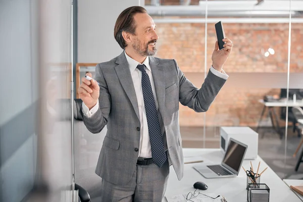 Cheerful man using cellphone and writing on whiteboard at work — Stock Photo, Image