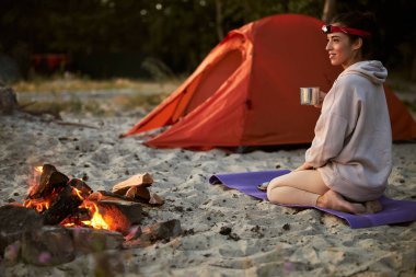 Kamp ateşinin yanında oturmuş çay içen neşeli genç bir kadın.