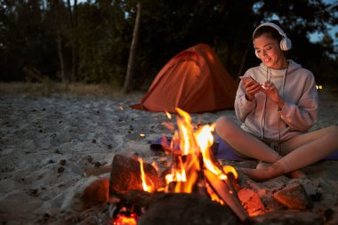 Neşeli genç bir kadın kamp ateşi yakarken cep telefonu kullanıyor.