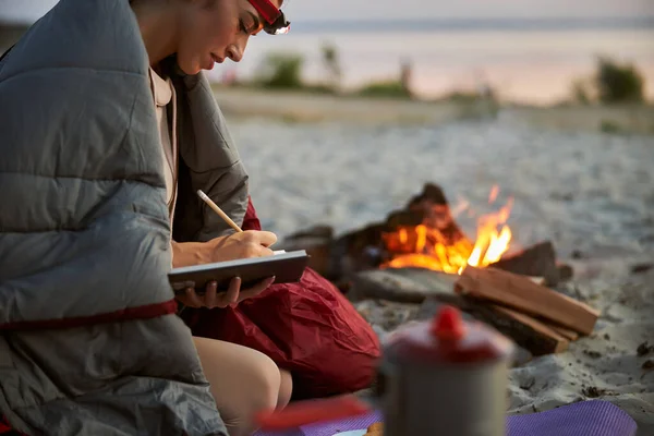 Charmante junge Frau schreibt in Notizbuch, während sie am Lagerfeuer ruht — Stockfoto
