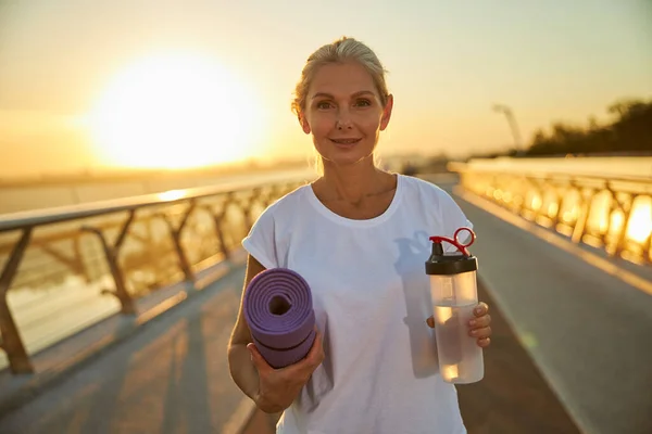 Mujer encantadora sosteniendo esterilla de yoga y botella de agua — Foto de Stock