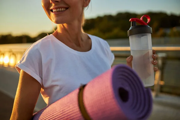 Mujer alegre sosteniendo esterilla de yoga y botella de agua — Foto de Stock