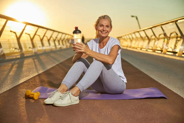 Mujer alegre y deportiva sentada en una esterilla de yoga al aire libre — Foto de Stock
