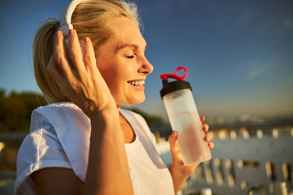 Mulher alegre em fones de ouvido sem fio segurando garrafa de água — Fotografia de Stock