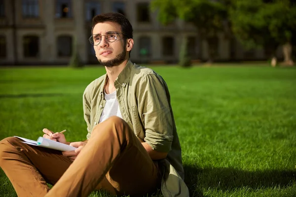 Gözlüklü düşünceli öğrenci çimlerin üzerinde oturuyor. — Stok fotoğraf