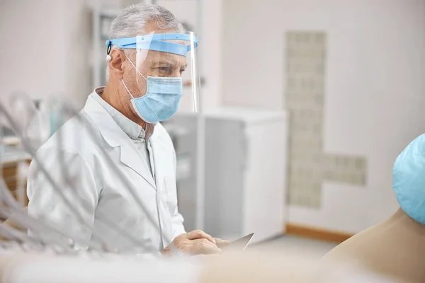 Uomo anziano che indossa uno scudo facciale con una tavoletta — Foto Stock